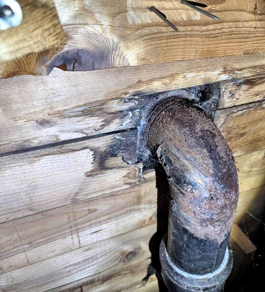 Rusted cast-iron closet bend and water-stained plank subfloor seen from below a leaking toilet