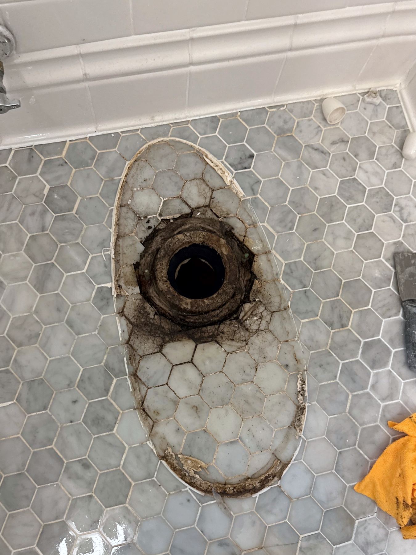 Toilet removed, showing a corroded flange, old wax seal and black staining spreading through the hex tile grout lines